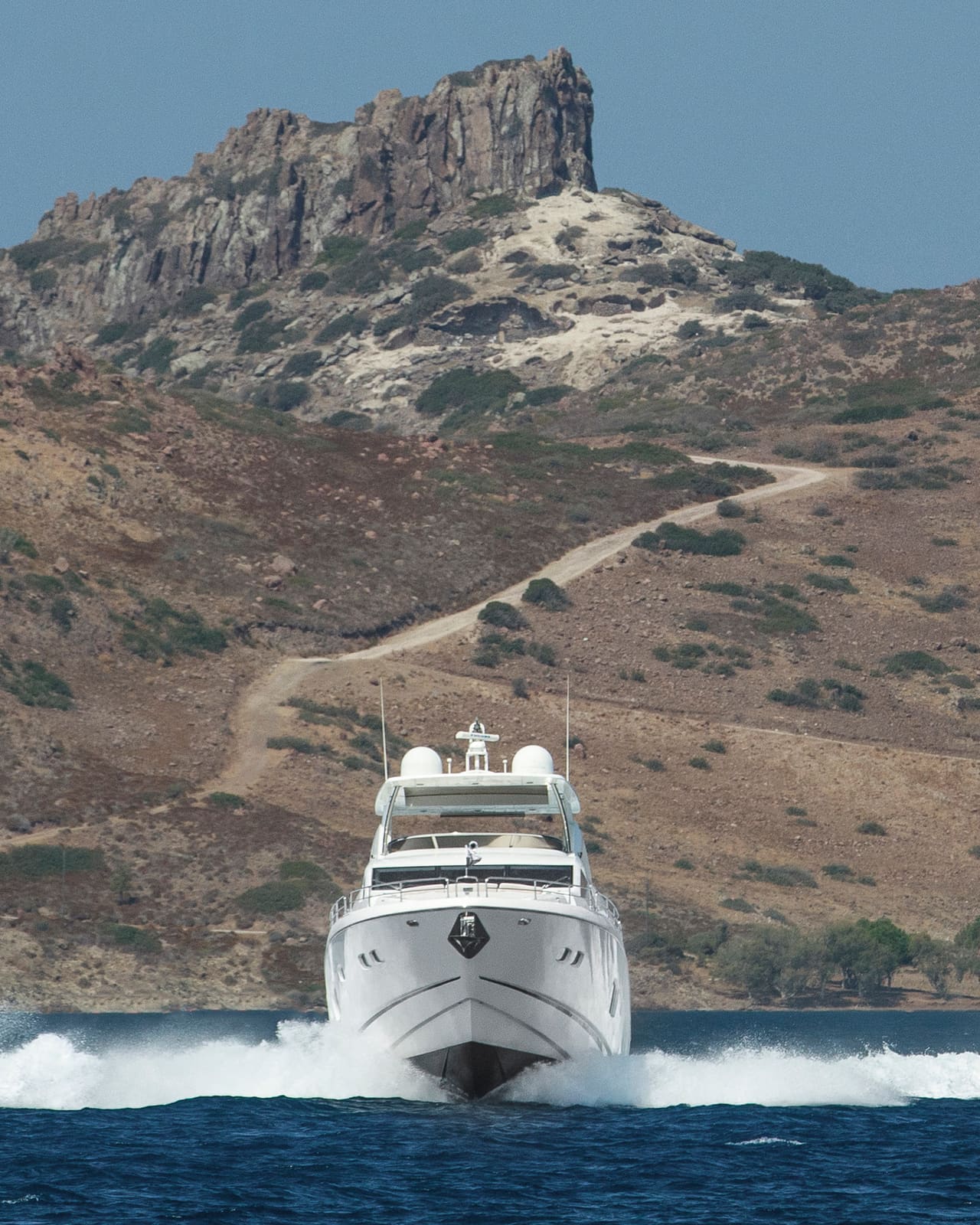 A motor yacht at speed, head-on, her bow wave white under a rocky hill
