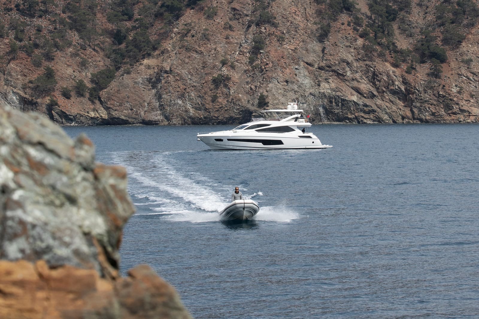 A motor yacht at anchor under a wooded hill, her tender crossing the bay